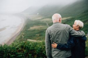 A senior aged couple embrace while looking out across a landscape
