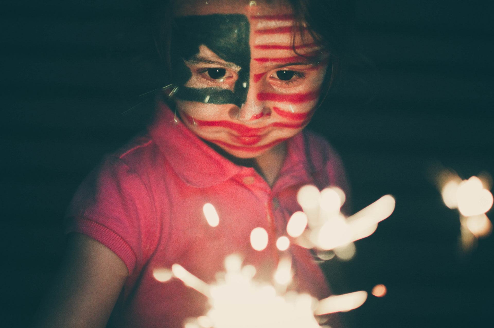 A young girl with her face painted red white and blue and a sparkler in her hand at night outside.